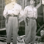 Dois homens vestindo uniforme, chapéu com aba e armas na cintura, olhando para o lado, enquadrados de corpo inteiro.