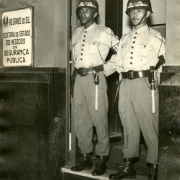 Dois brigadianos de uniforme e capacete, parados na entrada de uma porta aberta, enquadrados de corpo inteiro