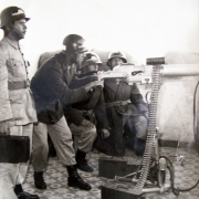 Oficial da brigada posicionado com metralhadora, junto a um grupo de outros três Oficiais, todos uniformizados com capacete.