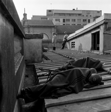 dois homens da brigada descansando em seus postos, aparentemente dormindo, cobertos por cobertor, em cima de um telhado. No fundo outros outros dois brigadianos uniformizados de pé.
