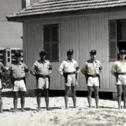 Grupo de oito oficiais da brigada uniformizados vestindo shorts e cap, posicionados lado a lado, em frente a uma casa.