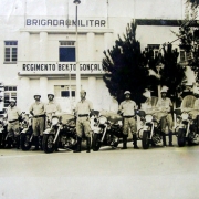 Grupo de homens uniformizados de capacete ao lado de suas motos, em frente ao prédio do Regimento.